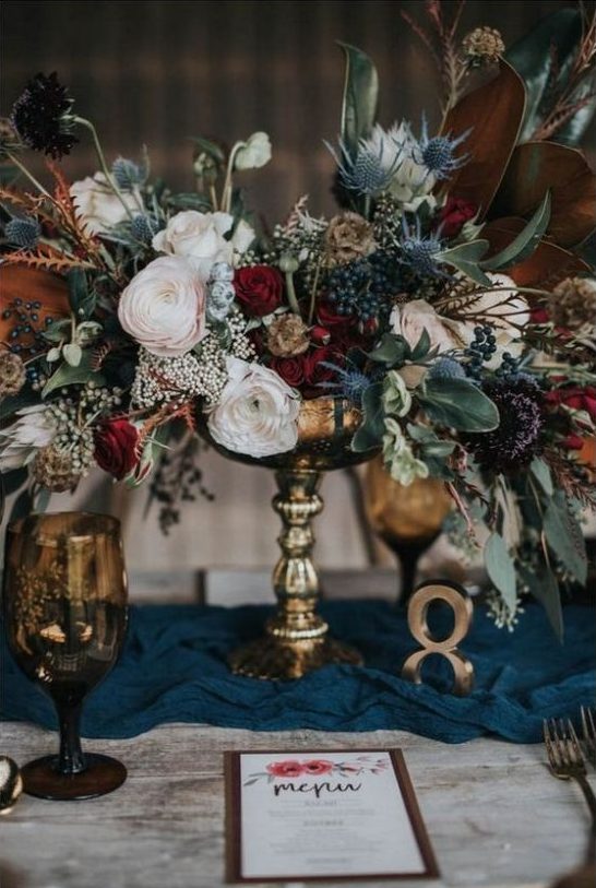 a stylish and textural winter floral centerpiece of burgundy, blush blooms, thistles, privet berries and foliage