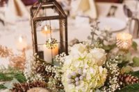 a vintage and rustic Christmas wedding centerpiece of greenery, fir branches, baby’s breath, white blooms and a vintage lantern with a candle