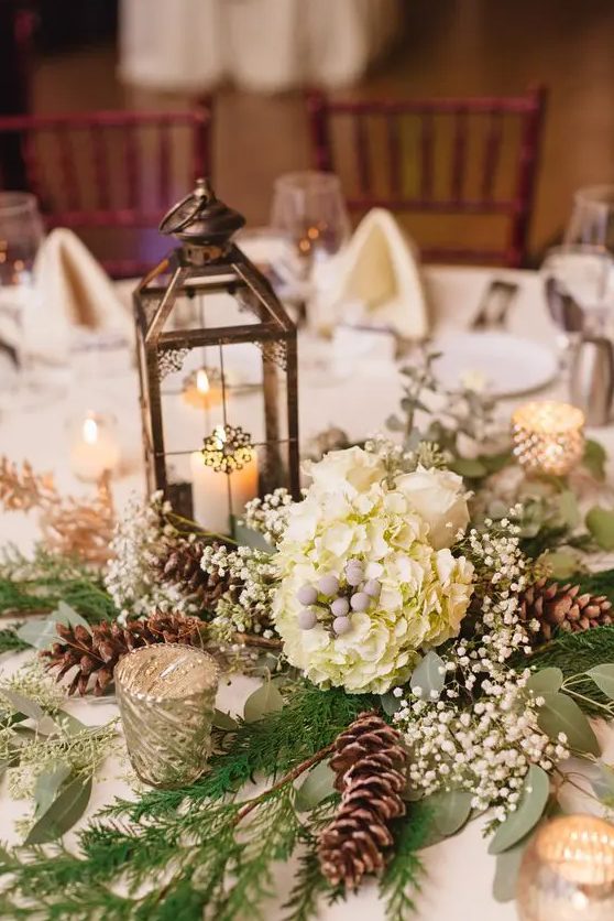 a vintage and rustic Christmas wedding centerpiece of greenery, fir branches, baby's breath, white blooms and a vintage lantern with a candle