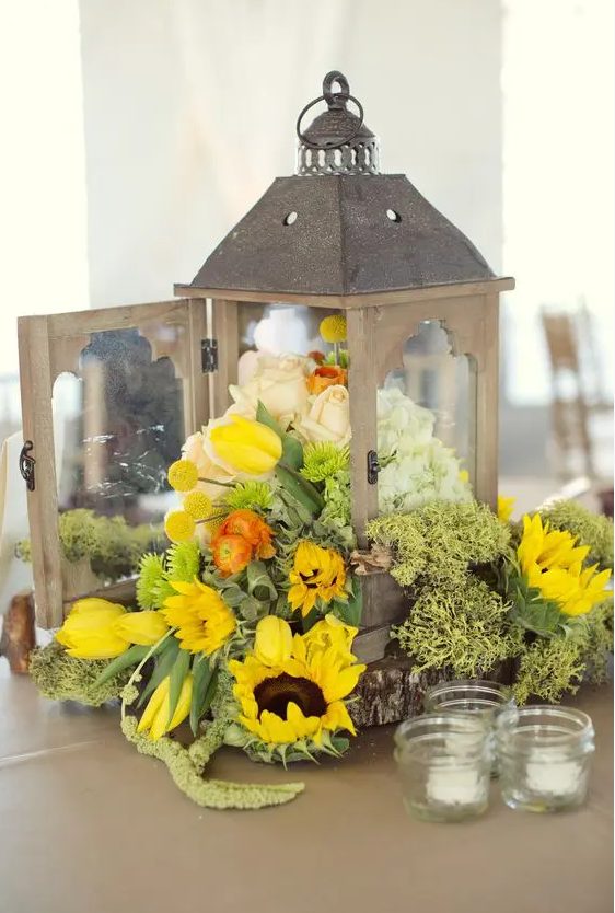 a vintage wedding centerpiece of a vintage candle lantern filled with sunflowers, yellow tulips, billy balls, white and blush roses and candles around