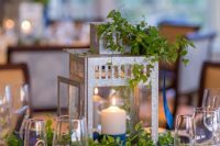 an eye-catchy wedding centerpiece of a lantern with a pillar candle, greenery and neutral blooms is a cool and fresh idea