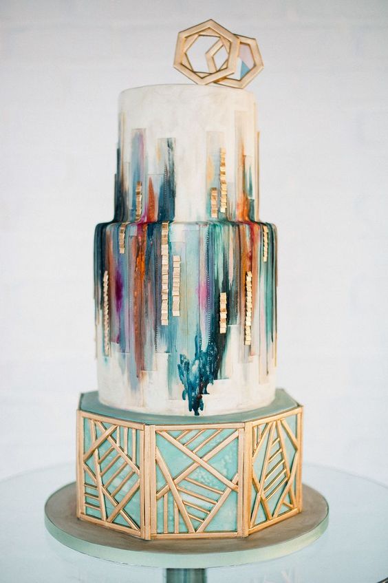 a wedding cake with colorful green, teal, orange, purple strokes, geometric gold decor and toppers