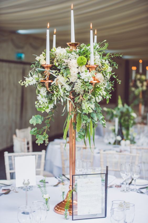 a tall copper candle stand with lush greenery and neutral flowers as a stunning centerpiece