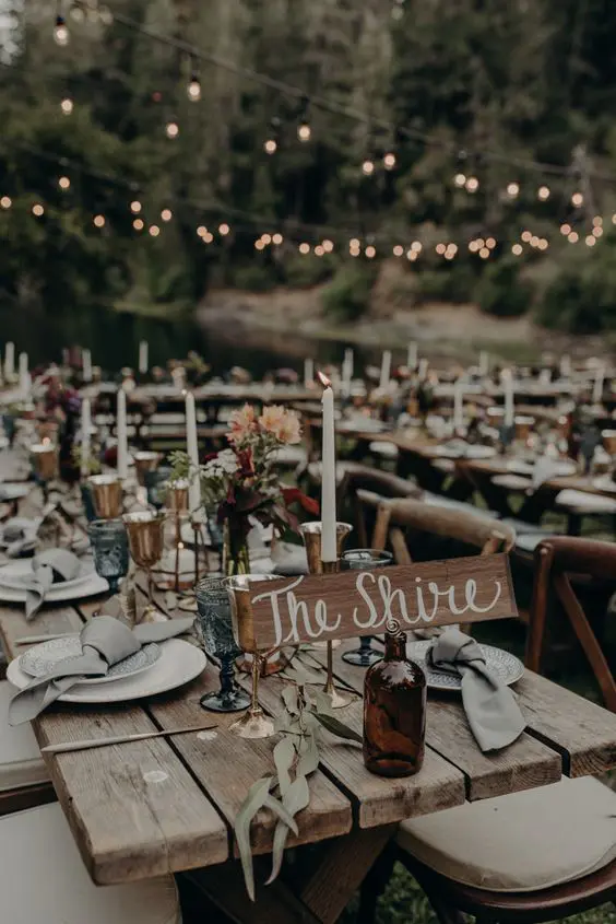 a LOTR wedding reception outdoors, with lights over the space, wooden tables and benches, greenery and blooms and candles