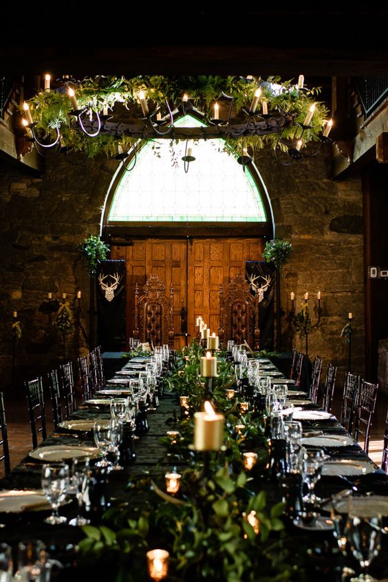 a LOTR wedding reception space indoors, with a black table with greenery and gold candles, a greenery chandelier with vines