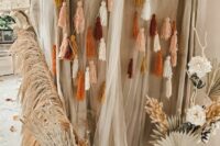 a boho photo booth wedding backdrop of neutral fabric and tulle, wooden beads and tassels, pampas grass and fronds is a lovely idea to rock