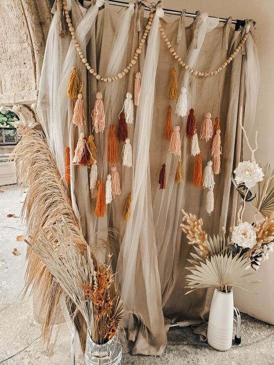 a boho photo booth wedding backdrop of neutral fabric and tulle, wooden beads and tassels, pampas grass and fronds is a lovely idea to rock