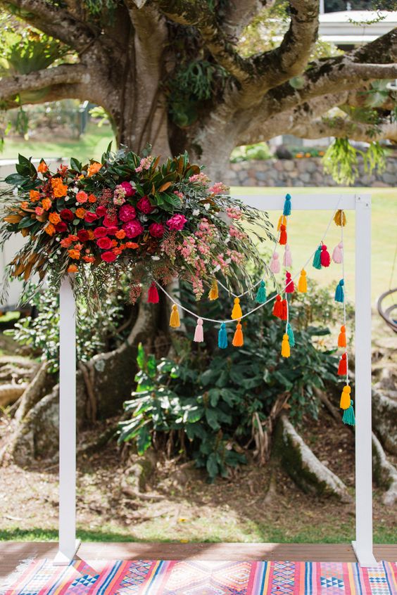 a colorful boho wedding backdrop with bright tassel garlands, bright fuchsia, orange and pink blooms and greenery for a Mexican wedding