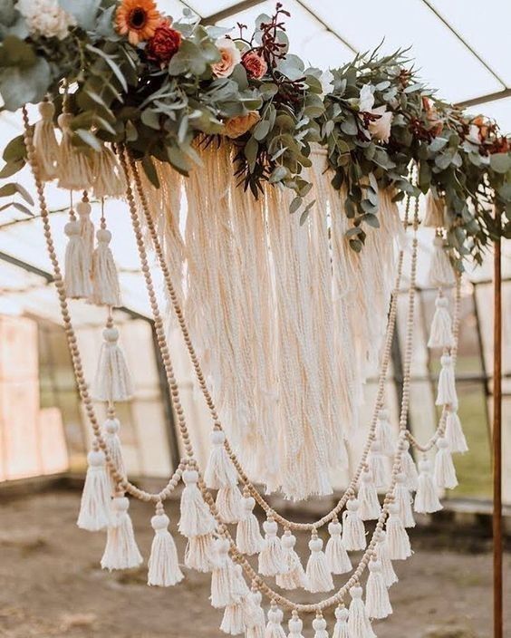 a creative boho wedding backdrop with long fringe, wooden beads and tassels, neutral and rust-colored blooms and greenery is super cool