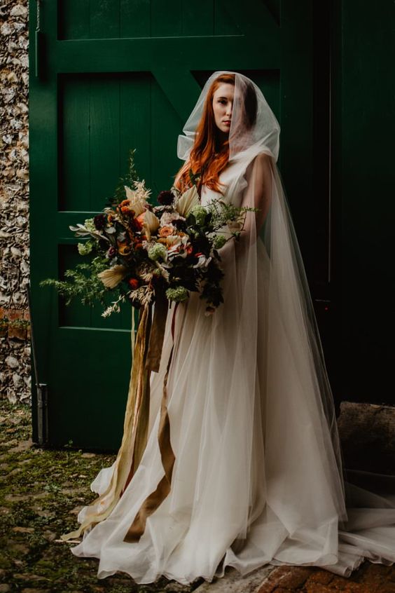 a fantasy bride wearing an A-line wedding dress, a sheer tulle capelet with a hood, carrying a lush wedding bouquet