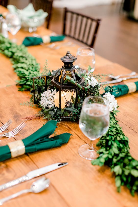 a lovely LOTR wedding table decor with greenery, a candle lantern and white blooms, green napkins and silver cutlery