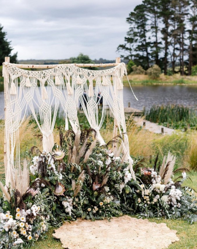 a macrame and tassel wedding backdrop with lots of greeneyr, white blooms and king proteas plus a rug is a great idea for a boho wedding