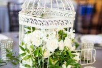 a neutral wedding centerpiece of a white cage with white blooms and greenery plus candles around