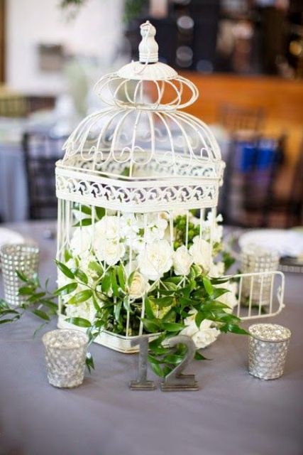 a neutral wedding centerpiece of a white cage with white blooms and greenery plus candles around