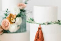 a plain white wedding cake decorated with greenery and berries and rust-colored tassels is a lovely idea for a bold modern wedding