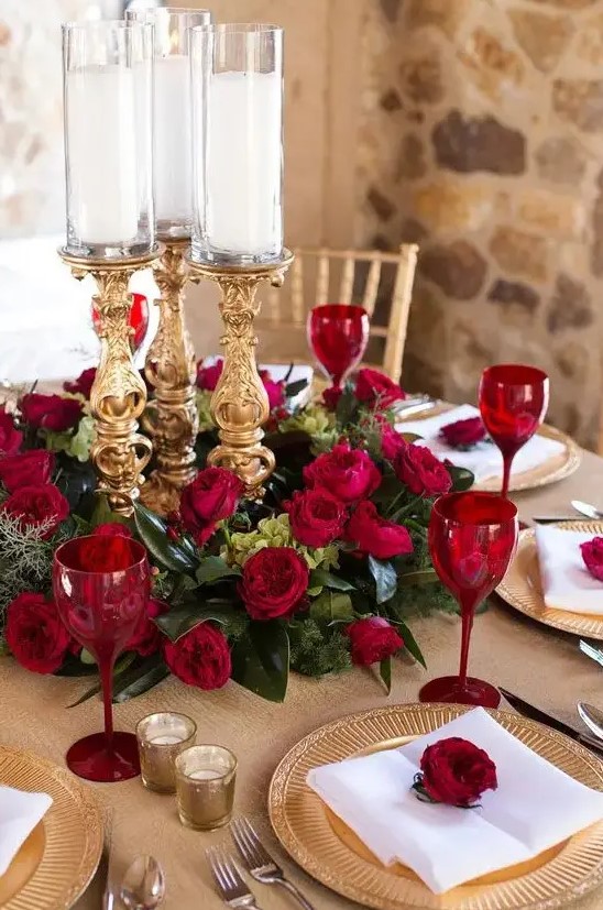a refined Valentine's Day wedding centerpiece of greenery, red roses, refined gold candleholders and red glasses to match