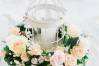a refined wedding centerpiece with a white cage with a bird on top and candle inside, surrounded with greenery and pink and peachy blooms