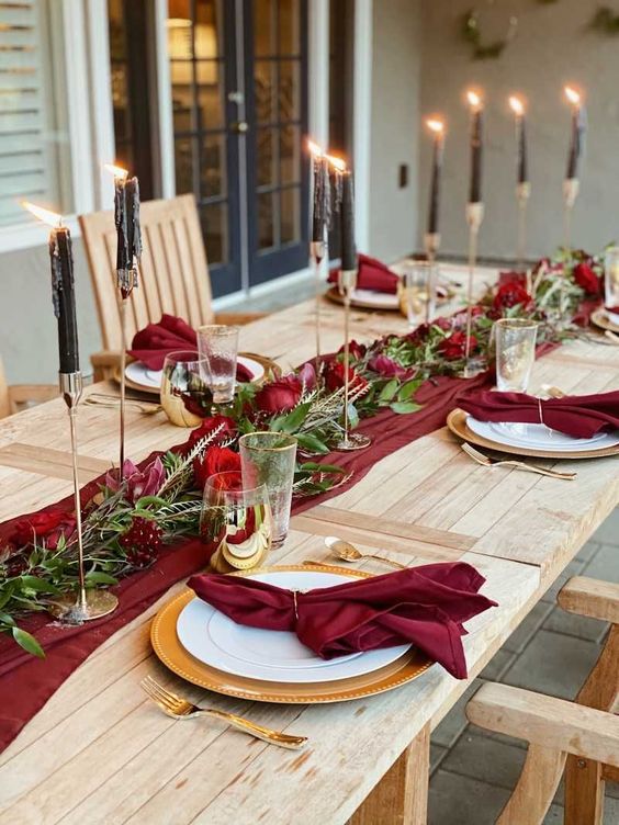 a stylish wedding table setting with a red runner and napkins, a greenery and red rose runner, black candles and white porcelain