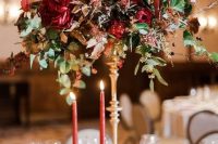 a tall wedding centerpiece with much texture, greenery, deep red and pink blooms and pink candles in gold candleholders