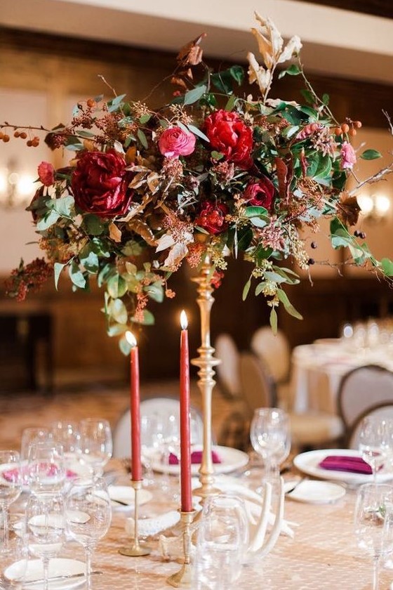 a tall wedding centerpiece with much texture, greenery, deep red and pink blooms and pink candles in gold candleholders