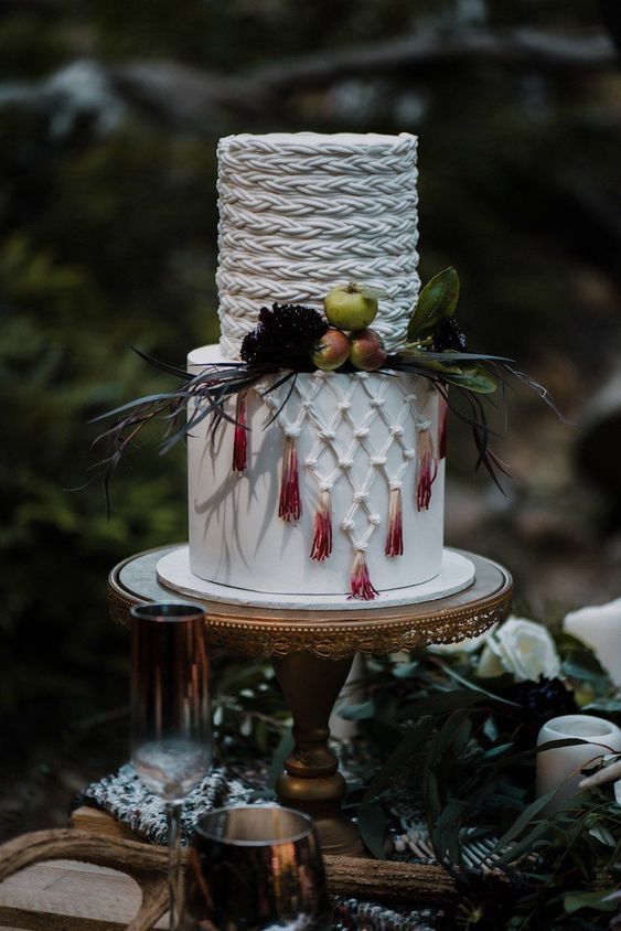 a white boho wedding cake with braids and macrame, with ombre tassels, fresh fruit and leaves is a lovely boho chic wedding idea
