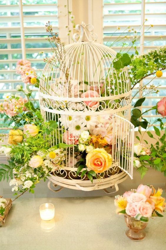 a white cage with white, blush and yellow blooms, blooming branches and greenery plus candles around