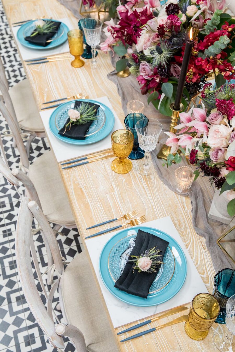 The tablescape features blue plates, blue and gold glasses, black candles in gold candle holders and various lush florals