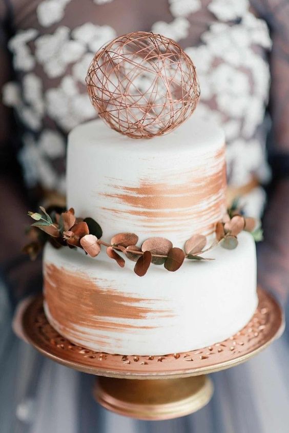 a white wedding cake decorated with copper, with a coppered greenery branch and a copper yarn ball on top