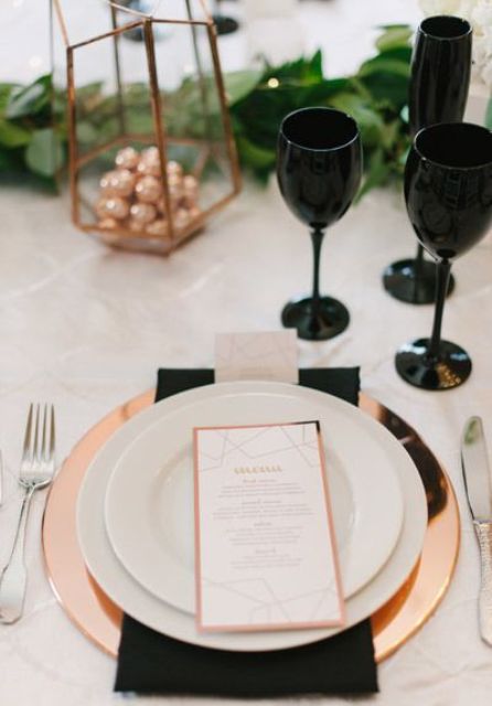 a modern glam place setting with a copper charger, black glasses, a copper lantern with candies and greenery