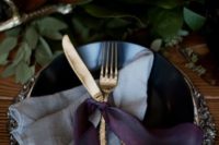 17 a moody place setting with a greenery table runner, dark plates and a silver charger