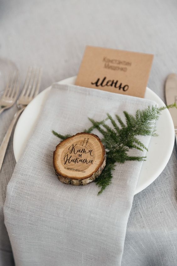 a cozy rustic place setting with a grey tablecloth and napkin, a wood slice, evergreens and a kraft paper menu