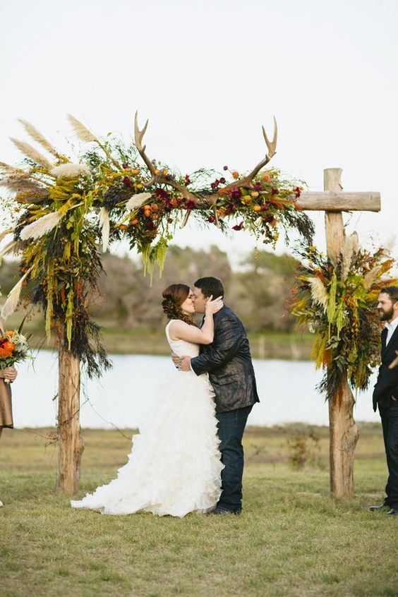 A boho wedding arch with lush greenery, pampas grass, fall colored blooms and antlers