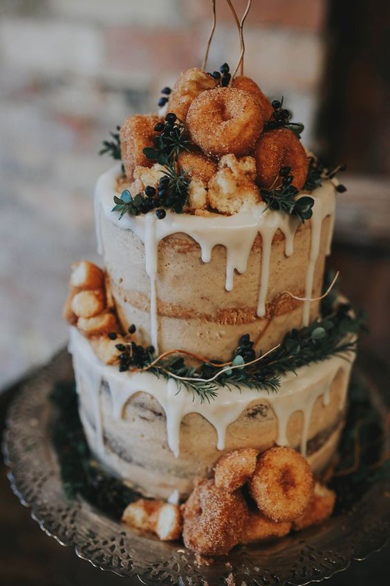 a naked wedding cake with dripping, evergreens, privet berries and topped with donuts