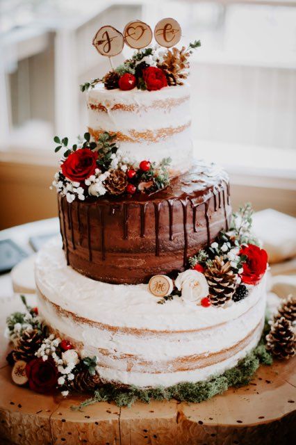 a Christmassy wedding cake with naked tiers and chocolate drip, red and white blooms, pinecones and wood slices