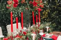 a bold Christmassy wedding tablescape with an evergreen and red rose table runner, a tall matching centerpiece, red roses and red napkins