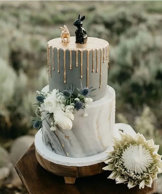 a bold wedding cake with a grey and grey marble tier, gold drip, white blooms, thistles and fun bunny cake toppers