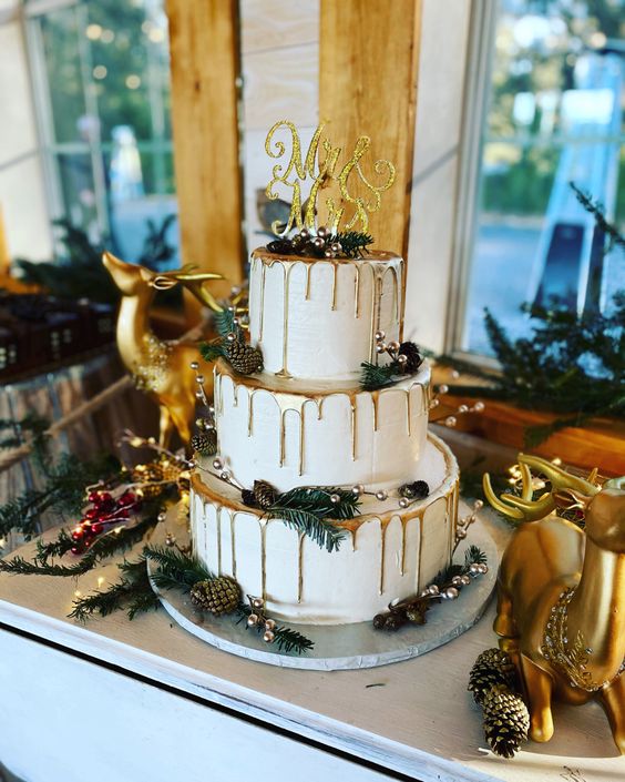 a catchy Christmas wedding cake with gold drip, evergreens, pinecones, berries and a gold glitter calligraphy topper