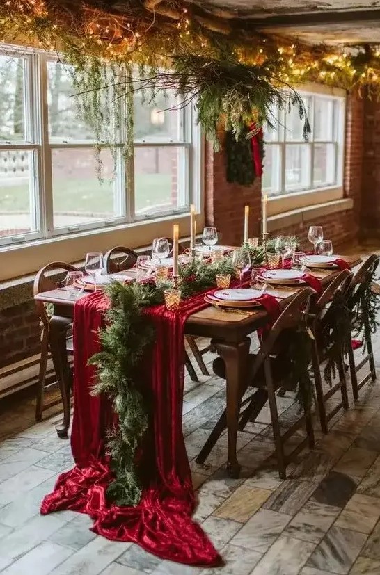 a lovely winter wedding reception space with evergreens and lights, a red table runner and evergreens and tall candles is chic