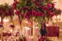 a lush Christmas wedding centerpiece of a tall gold stand with burgundy and red blooms and greneery plus candles around