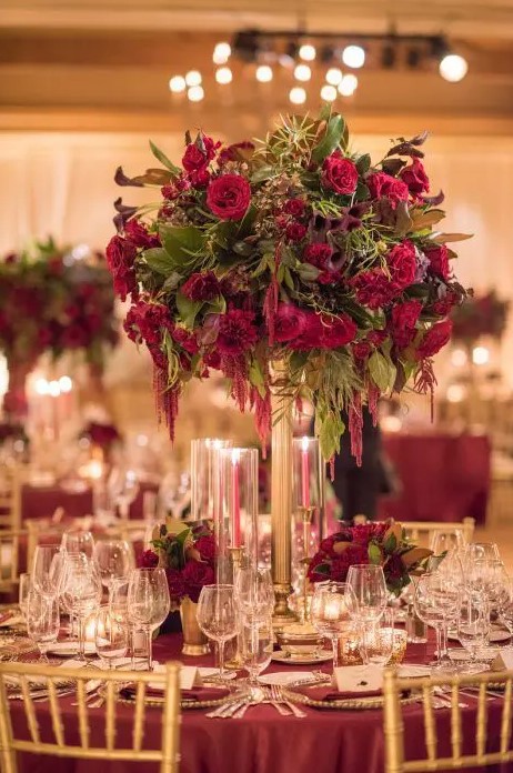 a lush Christmas wedding centerpiece of a tall gold stand with burgundy and red blooms and greneery plus candles around