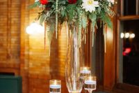 a refined Christmas wedding centerpiece of a tall glass vase with greenery, burgundy and white blooms, candles floating