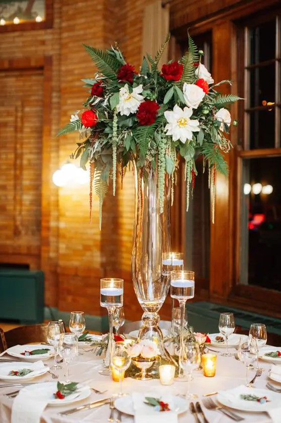 a refined Christmas wedding centerpiece of a tall glass vase with greenery, burgundy and white blooms, candles floating