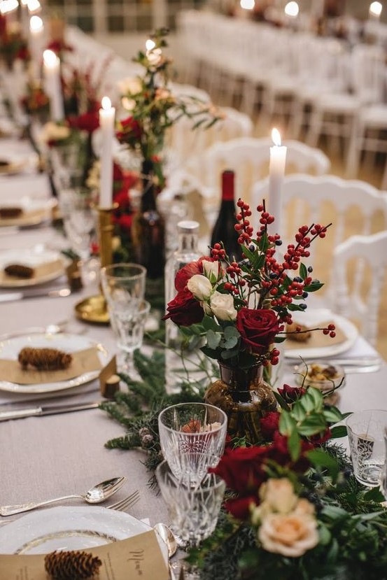 a traditional and chic Christmas tablescape with evergreens, greenery, white and red roses and berries, pinecones and tall and thin candles