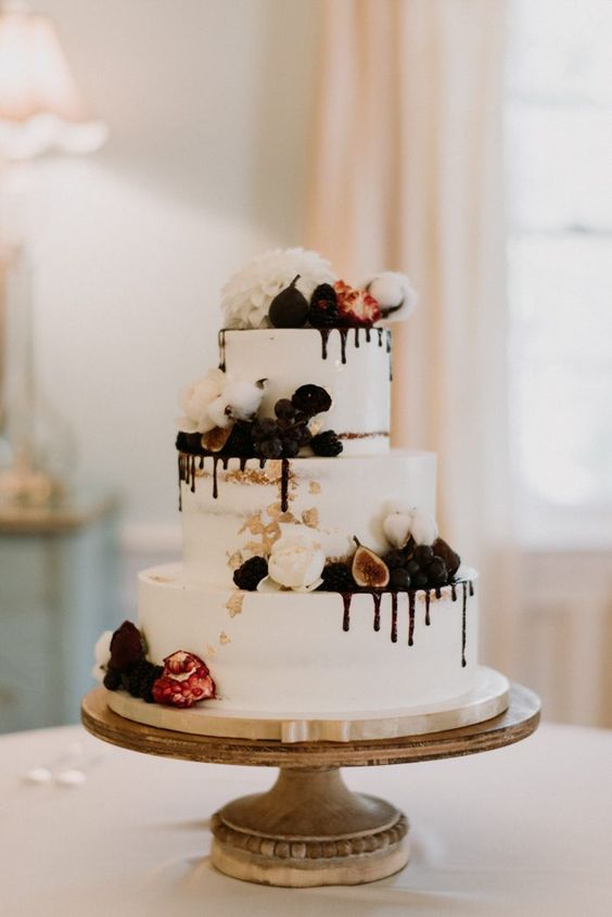 a white wedding cake with chocolate drip, cotton, figs and berries and white blooms on top is wow