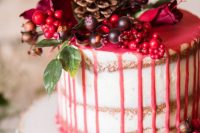 a winter wedding cake with pink drip, fresh berries and greenery and a pinecone on top is amazing
