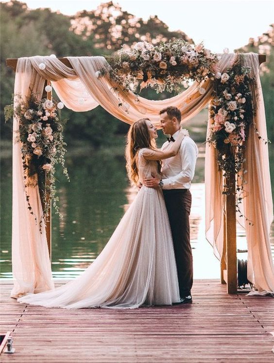 a pastel floral arch with blush and dusty pink blooms and blush draperies for a more romantic look