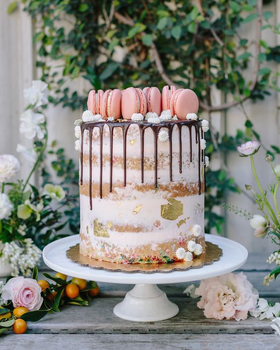 a naked cake with chocolate drip, meringues, pink macarons and gold leaf
