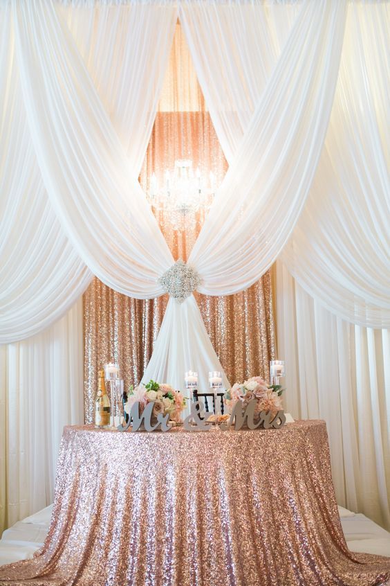 a rose gold sequin fabric backdrop plus white draperies, a large rhinestone brooch and a glam chandelier