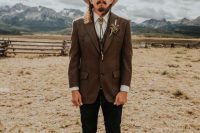 a boho meets rustic groom’s look with black jeans, a brown wiastcoat and blazer, amber shoes, a neutral hat and a bolo tie