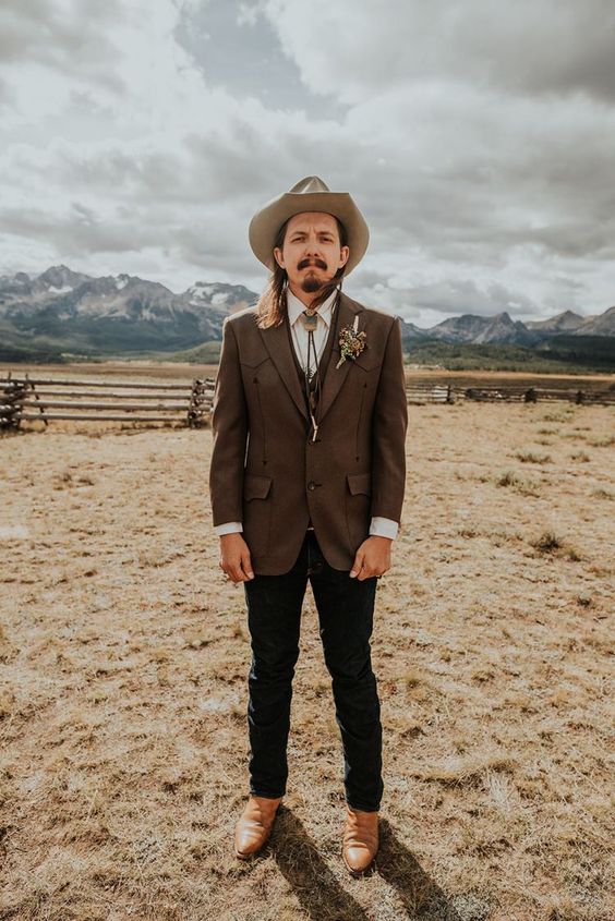 a boho meets rustic groom's look with black jeans, a brown wiastcoat and blazer, amber shoes, a neutral hat and a bolo tie